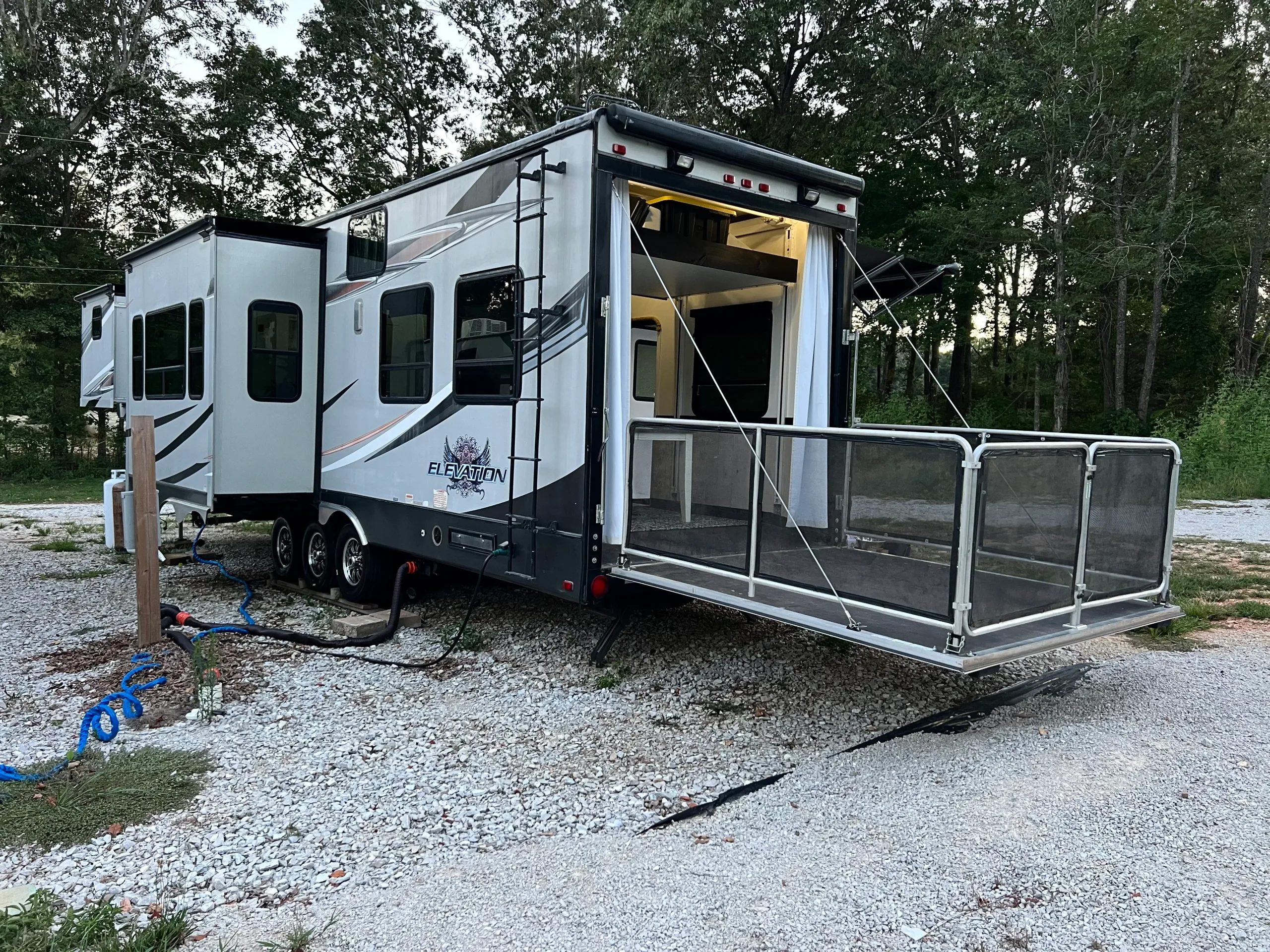 Toy hauler RV exterior with rear ramp set up as fenced patio and slide outs extended at wooded campsite.