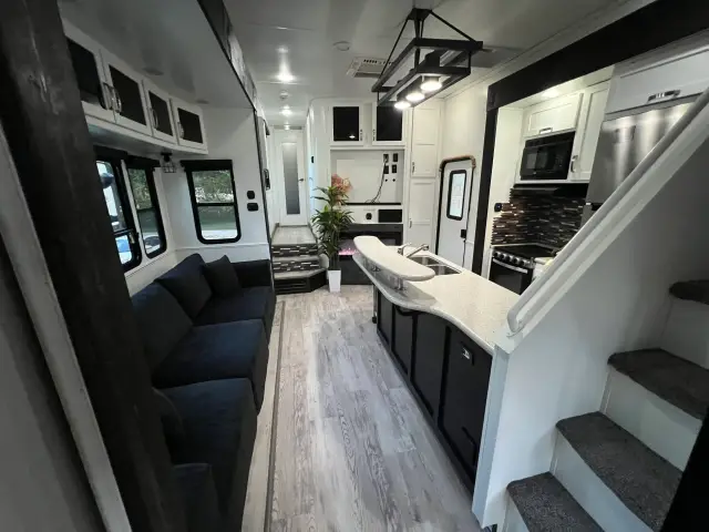 RV main living area with long sofa, kitchen island, stainless appliances, and steps leading to front hallway.