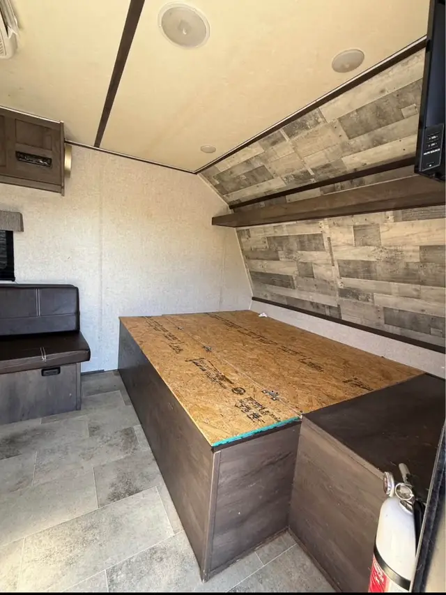 Interior front bedroom area with wooden bed platform, accent wall, and overhead shelf in a travel trailer.